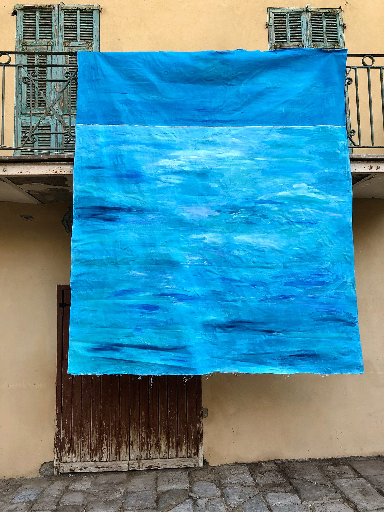 Large unstretched cotton painting in turquoise and deep blue hung across the ochre facade of a house in Bordighera Alta, above a wooden door and a wrought-iron balcony
